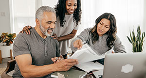 Ein älteres Paar und eine jüngere Frau sitzen gemeinsam an einem Tisch und gehen konzentriert Dokumente durch. Die jüngere Frau erklärt einen Punkt in den Unterlagen, während ein Laptop und eine Zimmerpflanze im Hintergrund zu sehen sind.