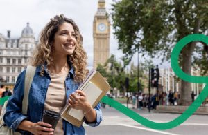 Eine junge Frau mit lockigem Haar hält in einer Hand einen Kaffeebecher und trägt in der anderen mehrere Bücher. Sie blickt sich fröhlich um, während im Hintergrund der berühmte Uhrturm Big Ben in London zu sehen ist.